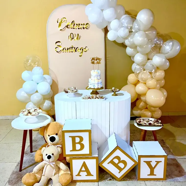 Mesa de bolo com arco de baloes brancos e dourados, ursinho de pelucia e cubos formando a palavra baby
